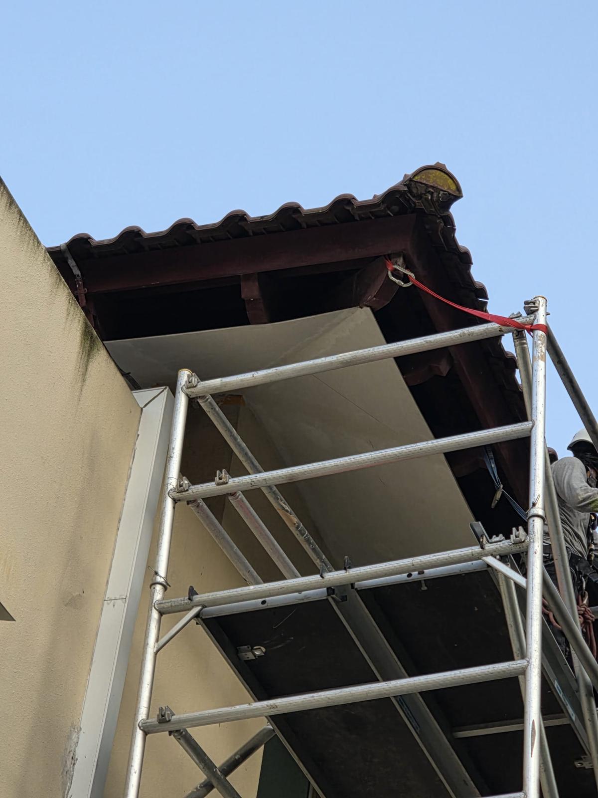 Worker in helmet and harness on a scaffold platform installing a waterproofed soffit board under the bungalow eave at Ewart Park second unit