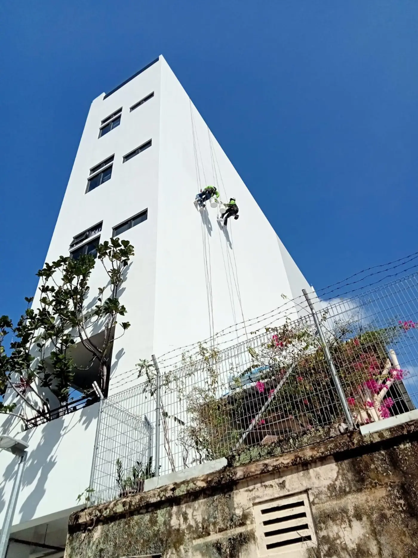 Two rope access technicians on twin ropes treating a spalled concrete edge on a high-rise HDB block facade in Bedok South, Singapore