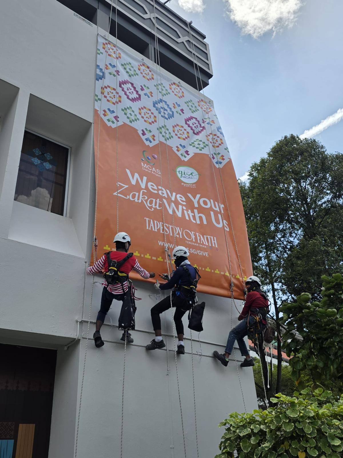 Three rope access technicians on twin ropes positioned beneath a newly installed MCAS Weave Your Zakat With Us – Tapestry of Faith banner on the building elevation