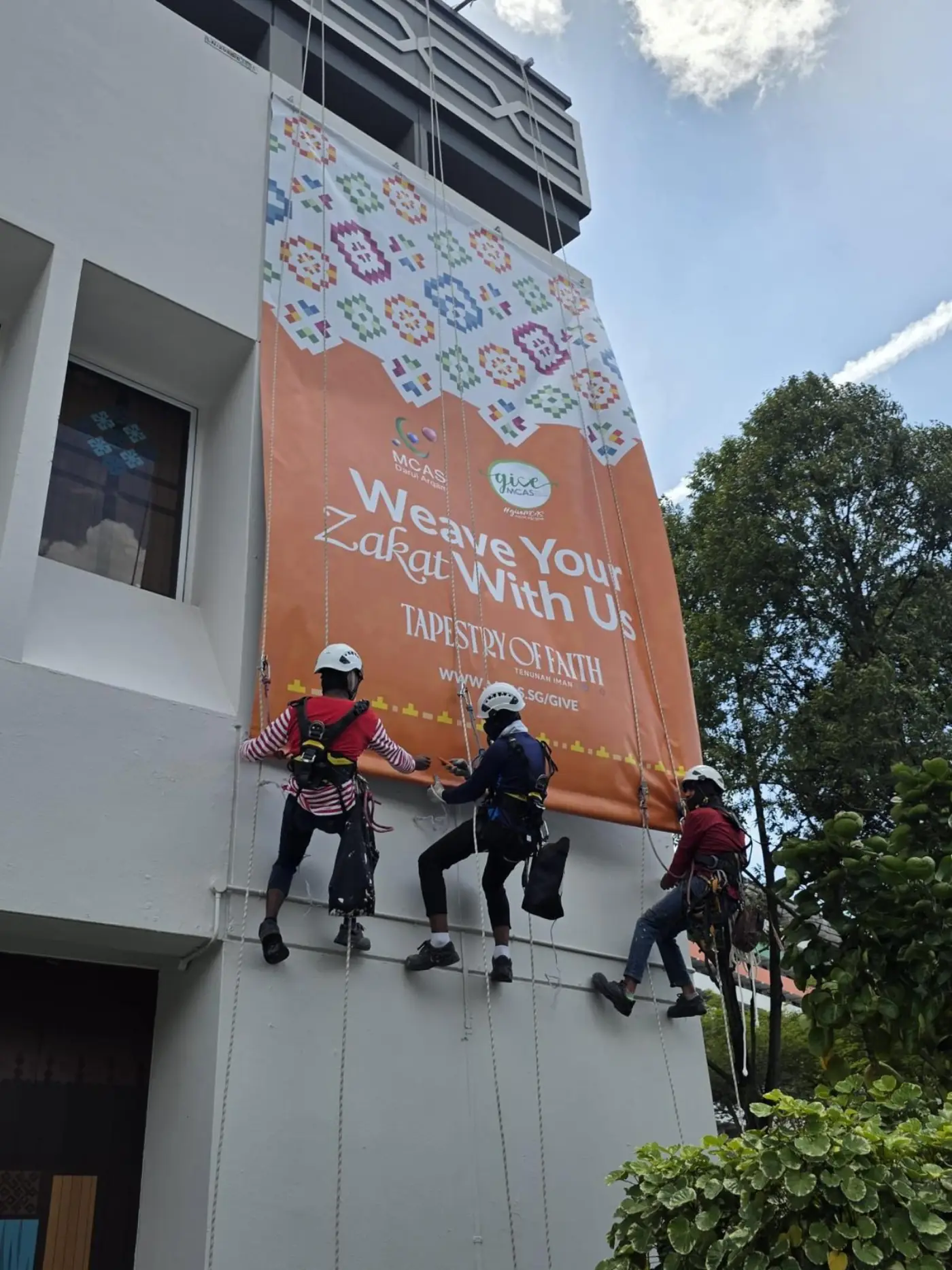 Three rope access technicians on twin ropes positioned beneath a newly installed MCAS Weave Your Zakat With Us – Tapestry of Faith banner on the building elevation