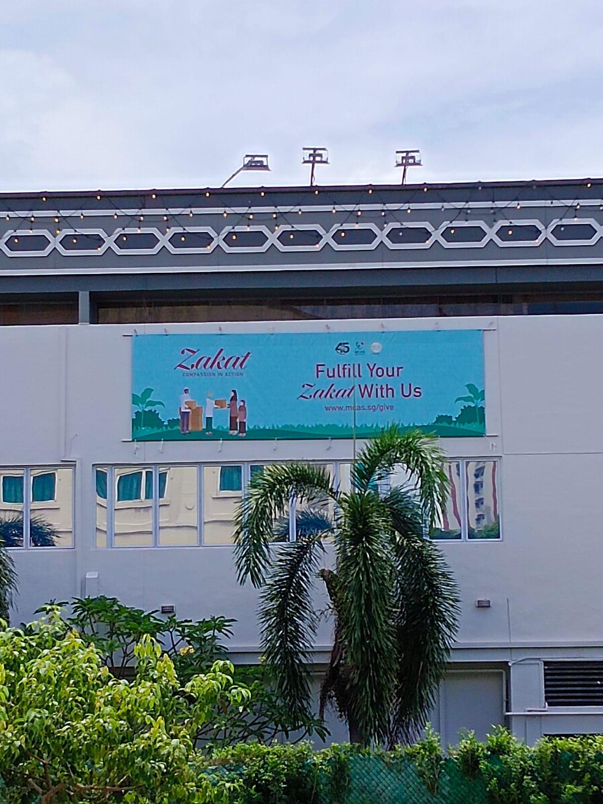 Secondary smaller Zakat banner installed on a neighbouring building elevation with a worker visible on the roof above