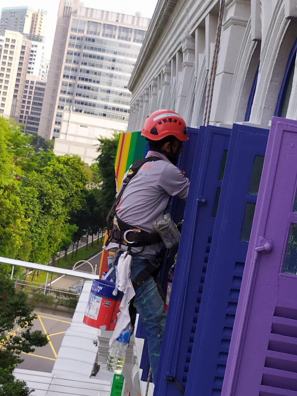 Rope access technician with tethered paint cans painting blue and green window shutters on the Old Hill Street Police Station facade