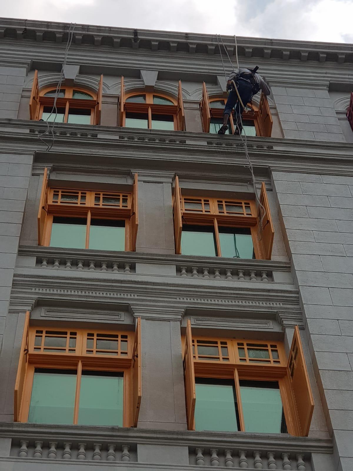 Rope access technician on a vertical descent line against the Old Hill Street Police Station facade between shutter bays during repainting