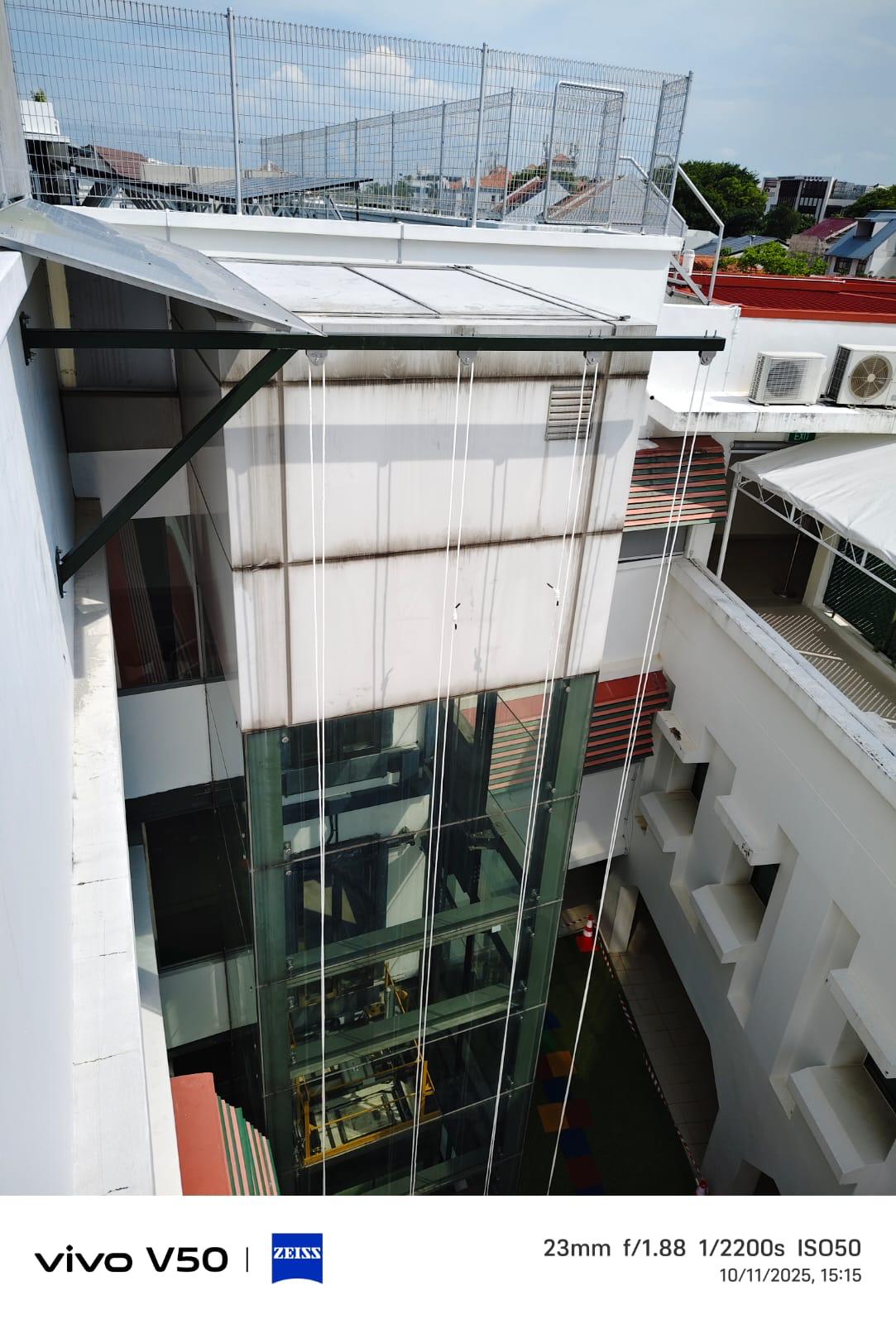 Full elevation view of a glass lift shaft with a black steel cantilever support bracket installed at the parapet and twin-rope access lines descending the face