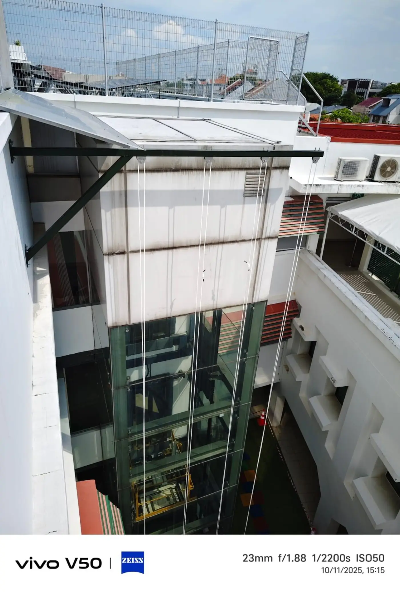 Full elevation view of a glass lift shaft with a black steel cantilever support bracket installed at the parapet and twin-rope access lines descending the face