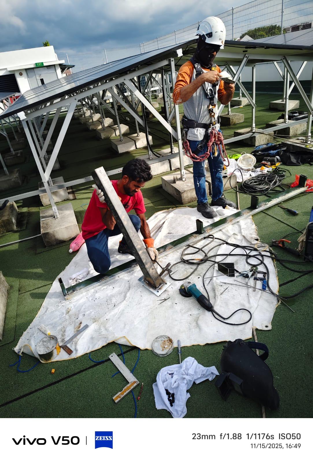 Two technicians fabricating steel support brackets on a rooftop solar deck; standing worker wears full-body harness and helmet with rope access PPE belt