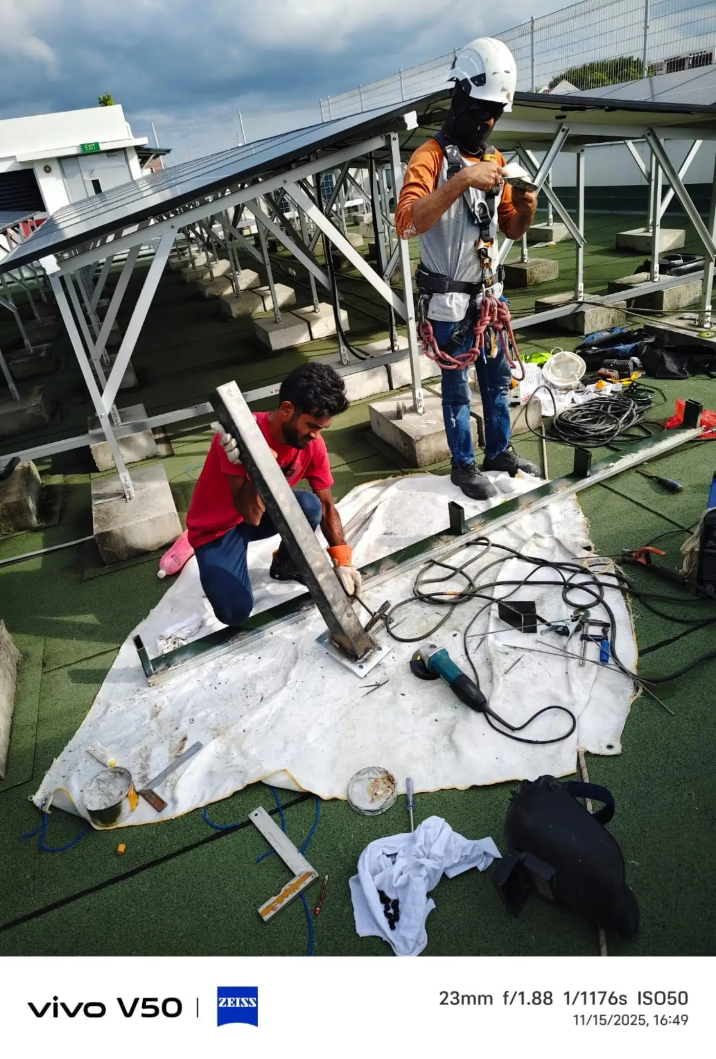 Two technicians fabricating steel support brackets on a rooftop solar deck; standing worker wears full-body harness and helmet with rope access PPE belt