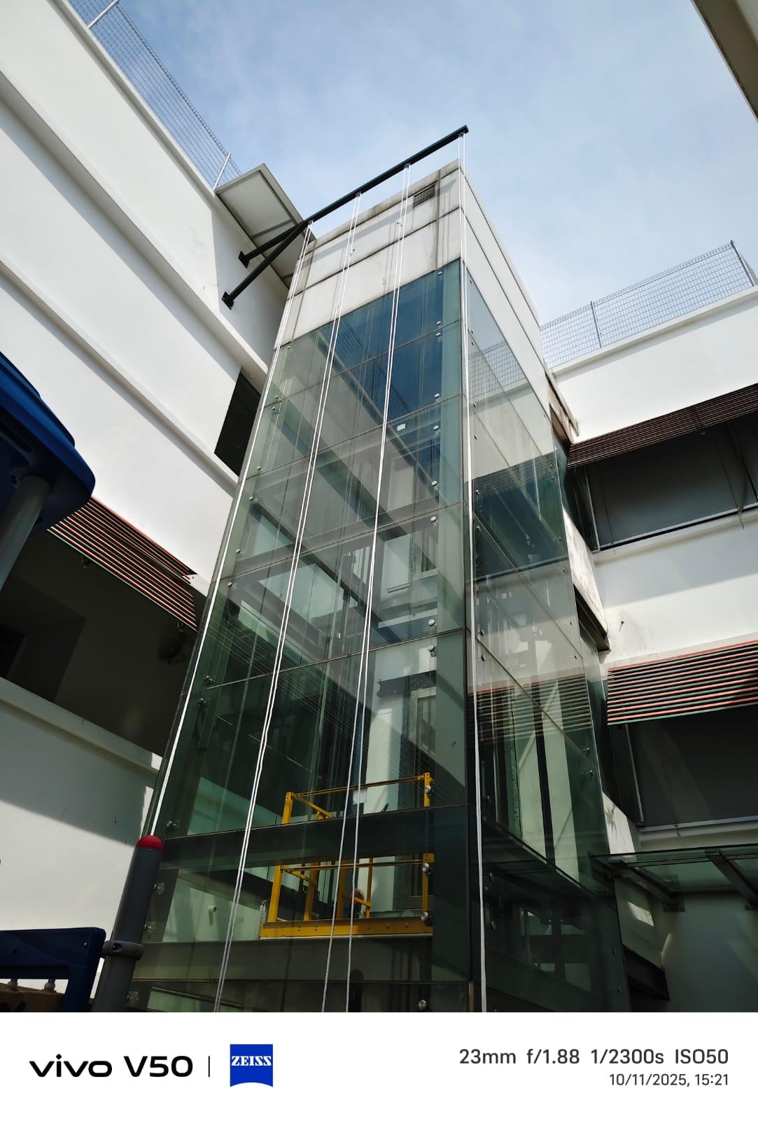 Detail view of the installed steel support beams at the top of the glass lift shaft with rope access lines descending past the elevation