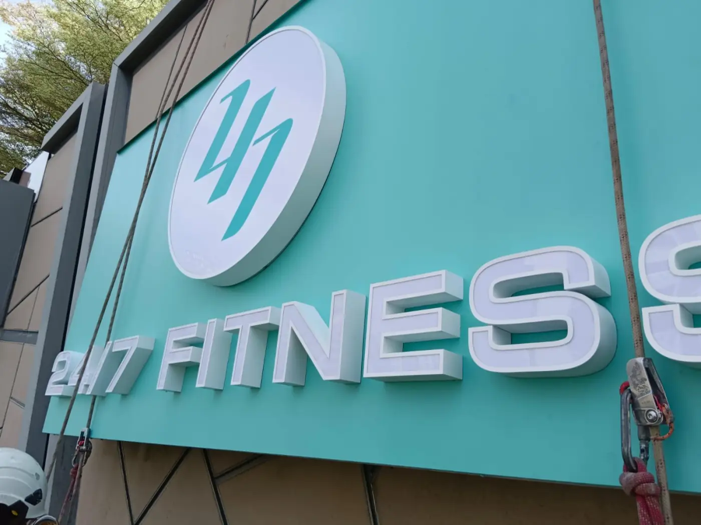 Close-up of installed fitness signage on The Seletar Mall tiled facade with rope access lines and a technician helmet visible at the lower frame